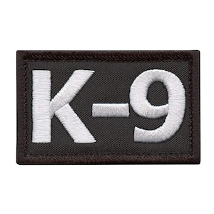Può includere: Toppa ricamata con la scritta "K-9" in filo bianco su sfondo nero. La toppa ha una forma rettangolare con un bordo nero. Le lettere sono in grassetto e facili da leggere.
