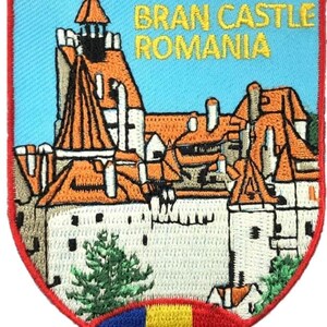 Può includere: Una toppa ricamata a forma di scudo raffigurante il Castello di Bran in Romania con tetti arancioni e mura bianche su un cielo azzurro. Il testo BRAN CASTLE ROMANIA è ricamato in giallo. La bandiera rumena è in basso con un bordo rosso.