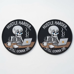 Peut inclure: Deux écussons brodés circulaires représentant un squelette blanc assis à un bureau avec un ordinateur portable et une tasse fumante. Le texte indique HUSTLE HARDER... STILL GONNA DIE. Le squelette a des larmes bleues et un cercle jaune.