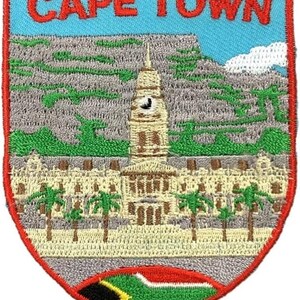 May include: A shield-shaped embroidered patch featuring the text CAPE TOWN in red letters. The design depicts a beige building with a clock tower, green palm trees, a grey mountain backdrop, and a small South African flag at the bottom.