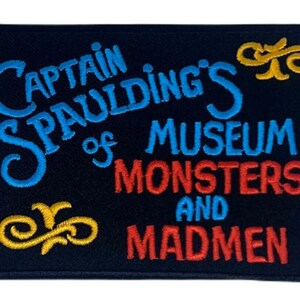 May include: A rectangular black embroidered patch featuring the text "CAPTAIN SPAULDING'S MUSEUM OF MONSTERS AND MADMEN" in blue and red thread, accented with two yellow decorative flourishes at the top right and bottom left corners.