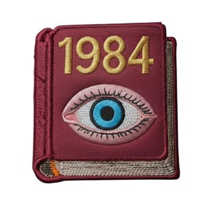Puede incluir: Parche bordado en forma de libro burdeos con el año "1984" en hilo dorado. La cubierta presenta un gran diseño de ojo en rosa, blanco y azul. El parche tiene un borde cosido.