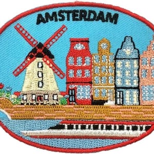 May include: An oval embroidered patch with a light blue background and a red border. It features a windmill, a row of colourful canal houses, and a boat on the water. The word AMSTERDAM is stitched in black letters at the top of the patch.