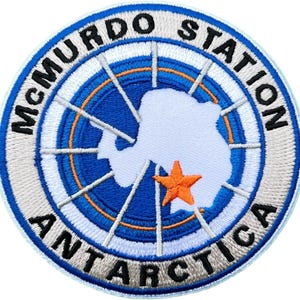 Op de afbeelding: Geborduurde patch met de tekst "McMurdo Station Antarctica" in een cirkelvormig ontwerp. De patch heeft een blauw en wit kleurenschema, met een oranje ster en een witte omtrek van Antarctica in het midden.