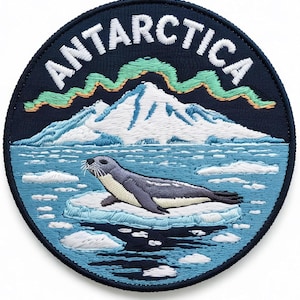 May include: A circular embroidered patch featuring a dark blue background with the word ANTARCTICA stitched in white at the top. The design depicts a grey seal resting on an ice floe in blue water, with a white snow-capped mountain and green aurora borealis.