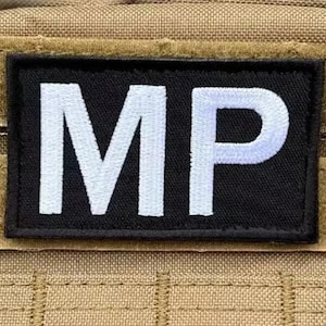Può includere: Una toppa rettangolare nera con le lettere bianche "MP" ricamate. La toppa è bordata di nero ed è attaccata a uno sfondo di tessuto color cachi.