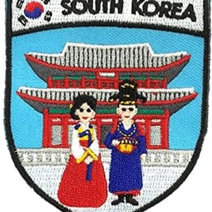 May include: A shield-shaped embroidered patch featuring the text SOUTH KOREA, a small South Korean flag, and a traditional building in red and grey. Two figures are depicted in the foreground wearing colourful traditional clothing on a light blue background.