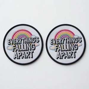 Könnte beinhalten: Zwei kreisförmige gestickte Aufnäher mit weißem Hintergrund und schwarzem Rand. Jeder Aufnäher zeigt einen bunten Regenbogen über hellblauen Wolken und den schwarzen Text "EVERYTHING'S FALLING APART" in einer fetten, serifenlosen Schrift.