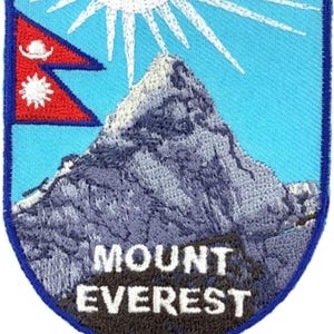 Könnte beinhalten: Ein schildförmiger, bestickter Aufnäher mit hellblauem Himmel, einer weißen Sonne, einem grauen Berggipfel und der Flagge Nepals in Rot, Blau und Weiß. Der Text MOUNT EVEREST ist in weißen Großbuchstaben am unteren Rand des Aufnähers eingestickt.