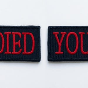 Op de afbeelding: Twee rechthoekige geborduurde patches met een zwarte stoffen achtergrond en de tekst YOU DIED gestikt in dikke rode hoofdletters. De patches hebben een nette, afgewerkte rand en liggen naast elkaar op een effen ondergrond.