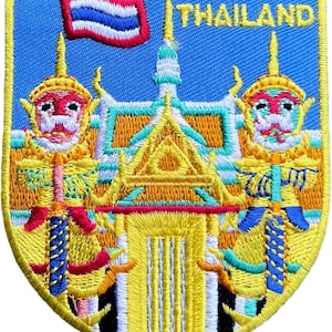 Puede incluir: Un parche bordado en forma de escudo con fondo azul y la palabra THAILAND en amarillo. El diseño incluye una estructura de templo amarilla central flanqueada por dos figuras guardianas rojas y doradas y una pequeña bandera tailandesa.