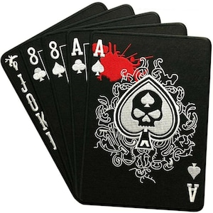 Könnte beinhalten: Ein Satz schwarzer Spielkarten mit weißen Stickereien. Die Karten zeigen die Zahlen 8, A und eine Joker-Karte. Das Pik-Ass hat ein Totenkopf-Design mit rotem Spritzer.