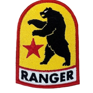 May include: An embroidered patch with a rounded top edge, featuring a black bear silhouette standing on its hind legs against a yellow background. A red five-pointed star is positioned to the left of the bear. A white horizontal bar at the bottom contains the black text RANGER, all bordered by a red stitched edge.