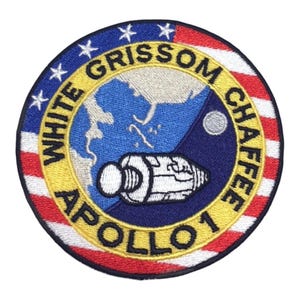 Pode incluir: Um emblema bordado circular com o emblema da missão Apollo 1. O design inclui uma representação da Terra, uma nave espacial e a Lua contra um fundo da bandeira americana. O texto diz WHITE GRISSOM CHAFFEE APOLLO 1.