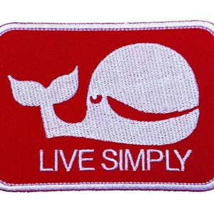 Könnte beinhalten: Ein rotes rechteckiges Patch mit einem weißen, gestickten Wal-Design und den Worten "LIVE SIMPLY" in Weiß. Der Patch hat einen weißen Rand und ist für Kleidung oder Accessoires bestimmt.