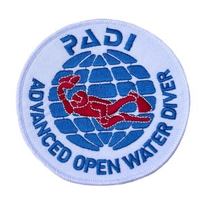 Puede incluir: Un parche bordado circular con fondo blanco y un gráfico de globo terráqueo azul. Una figura de buceador roja está centrada sobre el globo. El texto PADI está en la parte superior y ADVANCED OPEN WATER DIVER está escrito en azul en el borde.