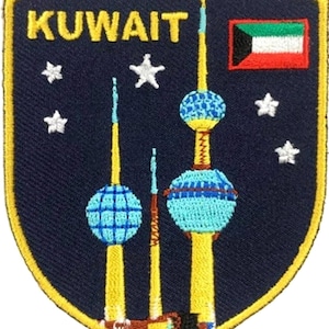 May include: A shield-shaped embroidered patch with a dark blue background, featuring the yellow text KUWAIT, a small Kuwaiti flag, and white stars. The design depicts three yellow towers with blue spherical sections against the dark blue field.