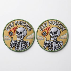 Peut inclure: Deux écussons brodés circulaires représentant un squelette blanc faisant un pouce levé sur un fond jaune. Au-dessus du squelette, le texte STAY POSITIVE est arqué en noir. Une météorite en feu est visible à gauche, avec des nuages sur les côtés.