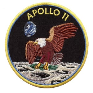 May include: A circular embroidered patch featuring a black background with the text APOLLO 11 in yellow. A bald eagle with brown and white feathers descends towards a grey lunar surface, carrying an olive branch. A blue and white Earth is visible.