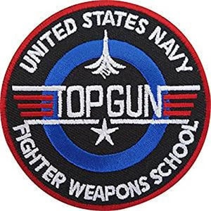Pode incluir: Um patch circular com o texto "UNITED STATES NAVY" e "TOPGUN FIGHTER WEAPONS SCHOOL". O patch apresenta um gráfico de avião a jato, uma estrela e listras vermelhas e brancas. O patch tem um fundo preto com detalhes em azul e vermelho.