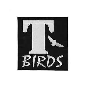 Puede incluir: Un parche cuadrado bordado en negro que presenta una letra T grande en blanco, la palabra BIRDS en letras mayúsculas blancas debajo y un pequeño pájaro blanco en vuelo posicionado a la derecha de la letra T, todo en un diseño cosido.