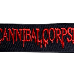 Puede incluir: Un parche rectangular bordado con fondo negro y el texto CANNIBAL CORPSE cosido en hilo rojo. Las letras presentan un efecto de goteo estilizado y todo el parche está bordeado por un fino borde negro cosido.