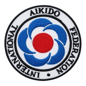 May include: A circular embroidered patch featuring a white background with a black border. The center displays a red circle surrounded by four blue curved shapes. The text "INTERNATIONAL AIKIDO FEDERATION" is stitched in black along the border.