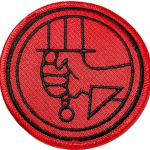 Può includere: Una toppa ricamata circolare con uno sfondo rosso vivace e un bordo nero spesso. Il design raffigura una grafica nera stilizzata di una mano che impugna una spada verticale, racchiusa all'interno di un sottile contorno circolare nero.