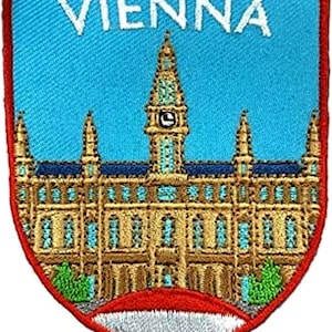 May include: A shield-shaped embroidered patch featuring a light blue background with the word VIENNA in white text. The design depicts a gold-colored building with a central clock tower, green accents, and a red and white striped base with a red border.