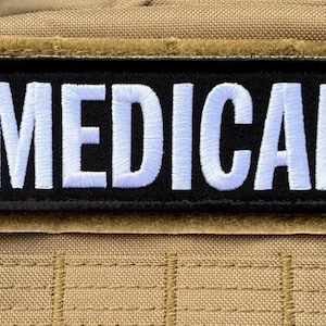 Puede incluir: Un parche rectangular negro con la palabra "MEDICAL" bordada en letras mayúsculas blancas. El parche está cosido sobre un fondo de tela color beige, posiblemente parte de un chaleco o bolsa táctica.