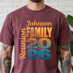 May include: A maroon short-sleeved t-shirt featuring a graphic print with the text Johnson FAMILY Reunion 2026. The design uses a gradient color scheme transitioning from yellow to orange to blue, with tree-like patterns integrated into the letters.
