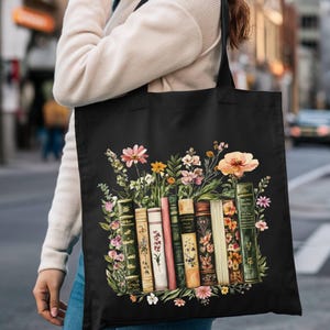 May include: Black tote bag featuring a colorful illustration of vintage books and flowers. The design includes a variety of book spines and a bouquet of pink, yellow, and orange flowers. The bag has black handles.