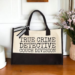 Two-tone canvas tote bag (beige and dark brown) with "TRUE CRIME DETECTIVE COUCH DIVISION" text, displayed on a wooden shelf next to a vase.