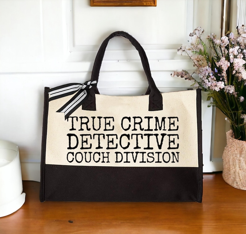 Two-tone canvas tote bag (beige and dark brown) with "TRUE CRIME DETECTIVE COUCH DIVISION" text, displayed on a wooden shelf next to a vase.