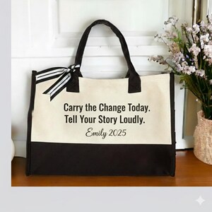 May include: A canvas tote bag with black handles and a black bottom panel. The bag has a cream-colored center panel with the text "Carry the Change Today. Tell Your Story Loudly. Emily 2025." A black and white striped bow adorns the side.