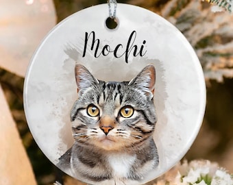 May include: A round, white ceramic ornament featuring a tabby cat portrait with the name "Mochi" in black script. The cat has yellow eyes and gray and brown stripes. A silver string is attached for hanging.