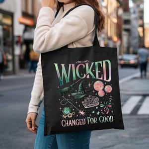 May include: Black tote bag featuring the words "WICKED" and "CHANGED FOR GOOD" in a colorful, gradient font. The design includes a witch's hat, broom, and other magical elements. The bag is carried by a person.