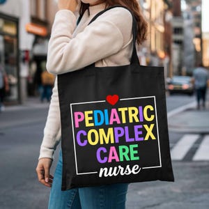 May include: Black tote bag with the words "PEDIATRIC COMPLEX CARE nurse" in colorful letters. A red heart is above the text. The bag is carried by a person in a cream-colored sweater and blue jeans.
