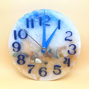May include: Round wall clock with a beach theme. The clock face features blue numerals and hands, with a sandy beach design and seashells. The top of the clock has a blue ocean design.