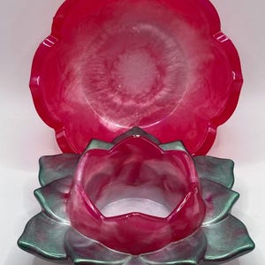 May include: A two-piece set of decorative resin containers. The top piece is a large, round, scalloped-edge dish in a vibrant pink hue. The bottom piece is a lotus flower-shaped container with pink and green metallic accents. The set is designed for decorative use.