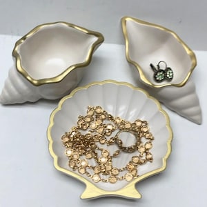 May include: Three white ceramic seashell-shaped trinket dishes with gold trim. One dish holds a gold necklace, and another holds a pair of green earrings. The dishes are ideal for storing jewelry or small items.