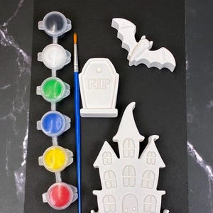 May include: A Halloween-themed craft kit featuring three white plaster figures: a bat, a tombstone with the text RIP, and a haunted house. The set includes a blue-handled paintbrush and a strip of six small paint pots containing black, white, green, blue, yellow, and red paint.