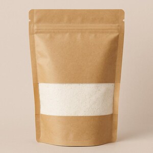 May include: A brown paper bag with a clear, horizontal window displaying a white, granular substance. The bag has a zip-top closure and is sealed. The background is a solid, light beige color.
