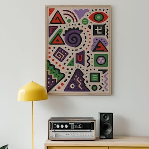 Op de afbeelding: Abstracte kunstprint in een houten lijst met kleurrijke geometrische vormen, waaronder driehoeken, spiralen en stippen. Het kunstwerk wordt tentoongesteld boven een stereosysteem en een gele lamp.