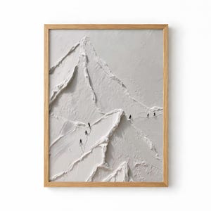 May include: A framed, textured white mountain art piece. The artwork depicts a mountain range with tiny figures climbing the slopes. The frame is made of light-coloured wood. The art piece is a minimalist, modern design.