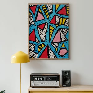 May include: Abstract art print with a geometric design. The artwork features red, pink, and yellow triangles against a blue background, with black dotted and lined accents. The artwork is framed in a light wood frame.