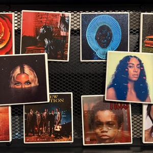May include: A collection of square ceramic coasters featuring album art from various musicians. The coasters showcase vibrant colors and portraits, including images of Stevie Wonder, Beyoncé, and Solange. The coasters are displayed in a black metal grid.