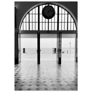 Könnte beinhalten: Schwarz-Weiß-Fotografie eines gewölbten Eingangs, der zu einem Strand führt. Über dem Eingang hängt eine runde Uhr. Der Boden hat ein geometrisches Fliesenmuster. Zwei Personen sind am Strand zu sehen.