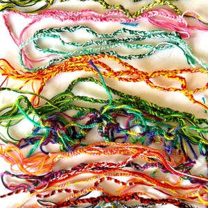 May include: A collection of colorful friendship bracelets in various shades of pink, green, yellow, orange, and blue. The bracelets are made of braided and twisted threads, with a handmade appearance. The background is a plain white surface.