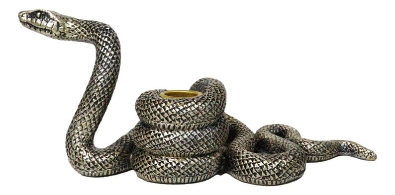 K&ouml;nnte beinhalten: Ein silberfarbener, schlangenf&ouml;rmiger Kerzenhalter. Der K&ouml;rper der Schlange ist gewunden, mit erhobenem Kopf und einem goldfarbenen Kerzenhalter in der Mitte. Die Schlange hat ein strukturiertes, schuppenartiges Muster.